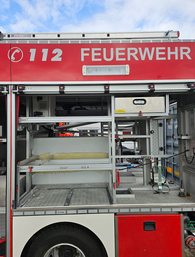 Ein rotes Feuerwehrauto mit offener Seitenklappe zeigt Regale und Ausrüstung.