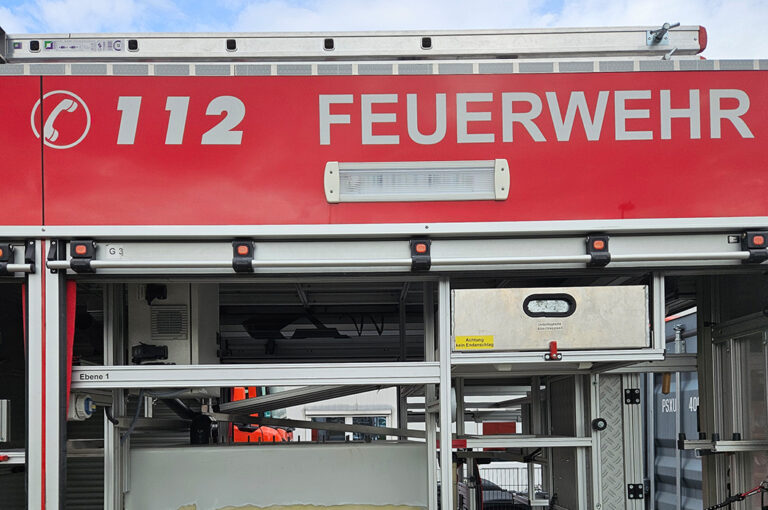 Ein rotes Feuerwehrauto mit offener Seitenklappe zeigt Regale und Ausrüstung.