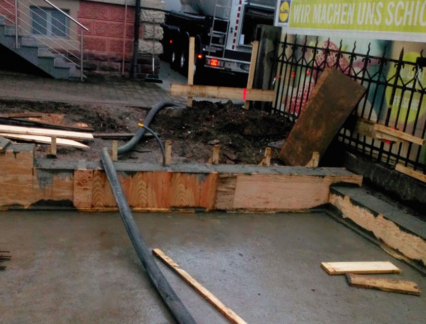 Eine Baustelle mit frischem Beton, Holzbrettern und dunklen Rohren bei trübem Licht