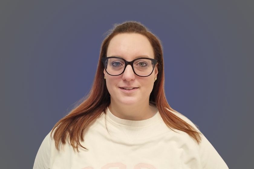 Frau mit langen rötlichen Haaren, Brille und hellem Shirt lächelt freundlich