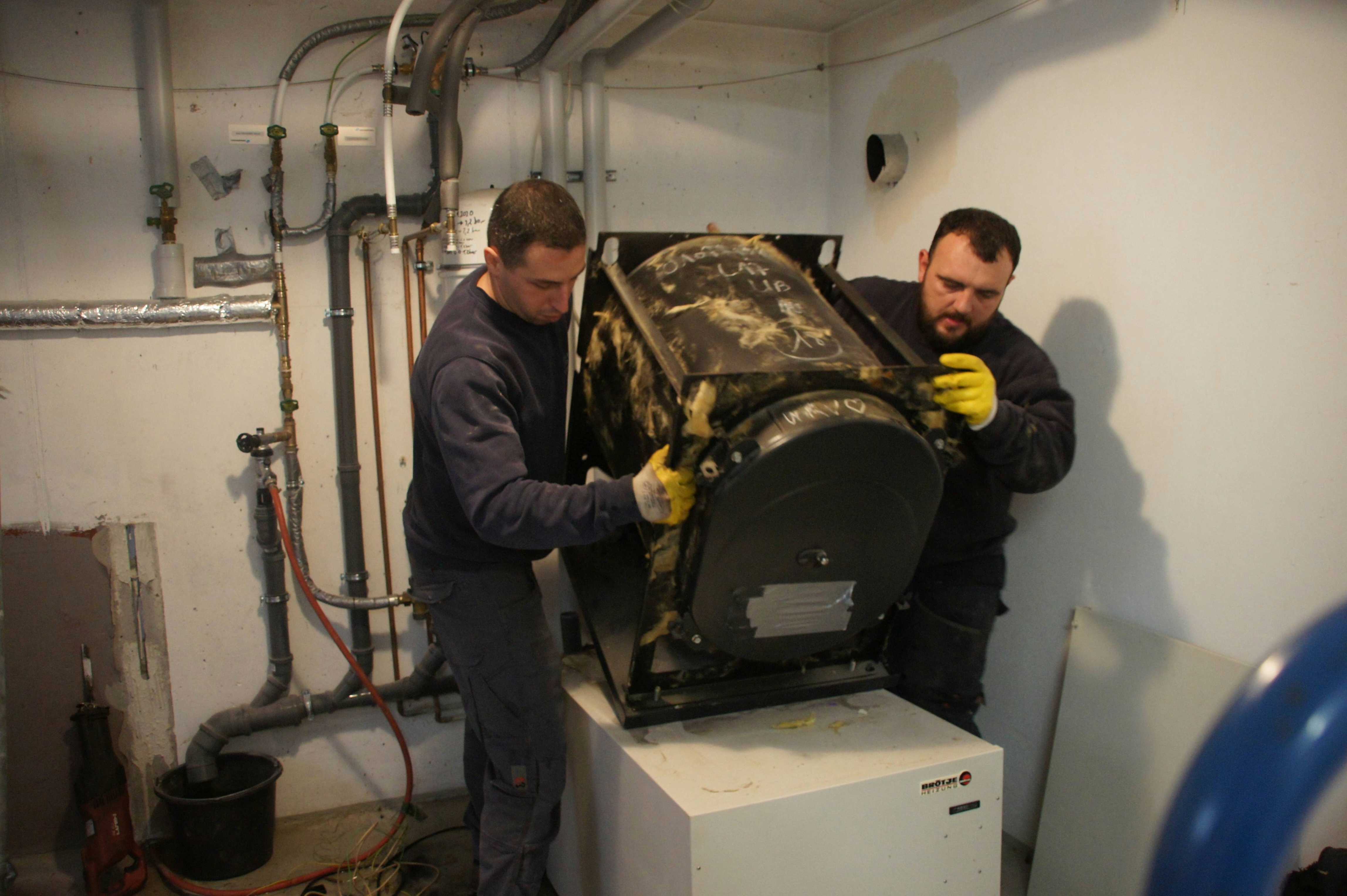 Zwei Männer mit konzentriertem Blick heben gemeinsam einen rußigen schwarzen Heizkessel